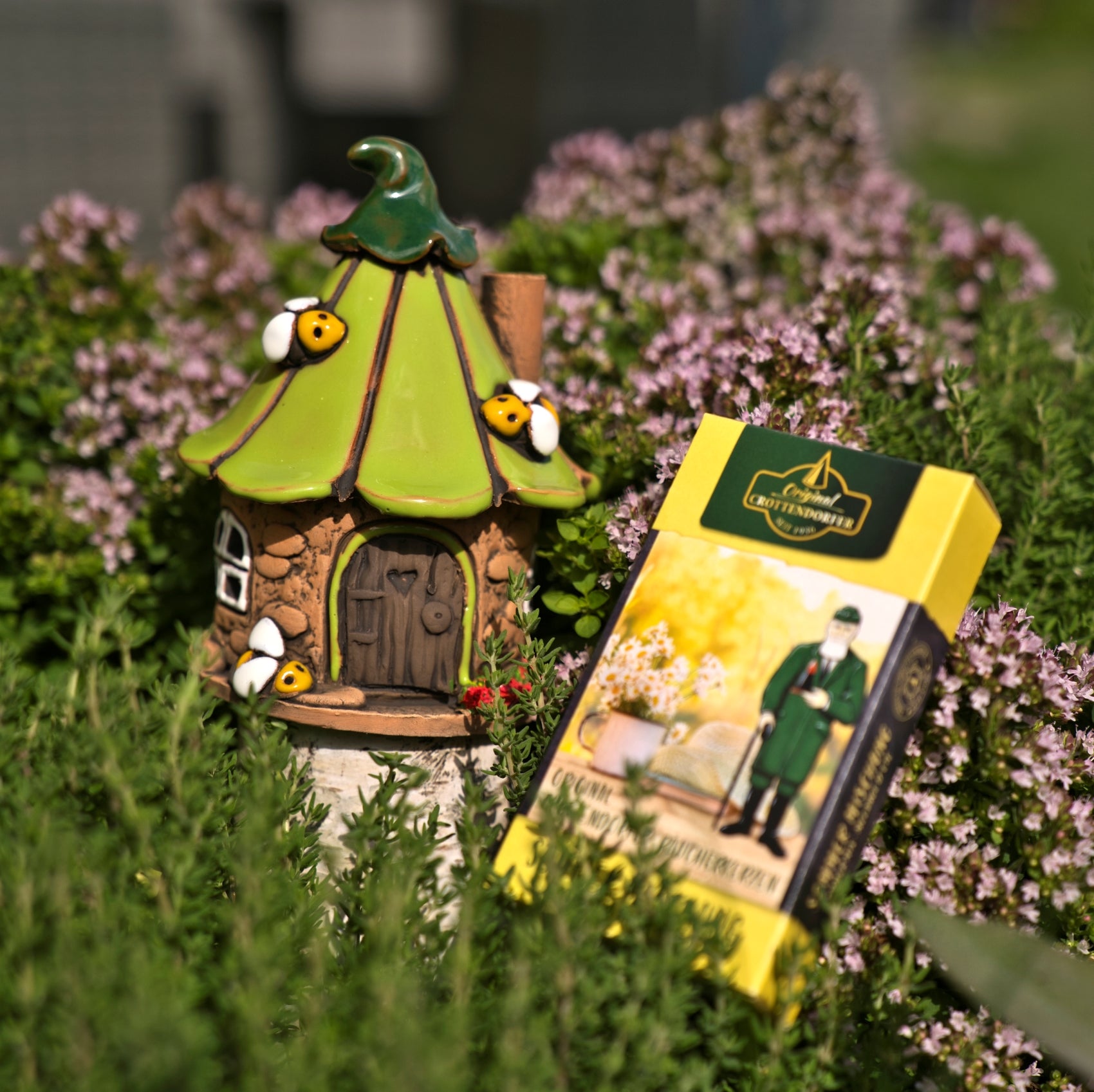 Das Original Crottendorfer Räucherhaus-Keramik-Bienenhaus, liebevoll gefertigt und mit einem dekorativen grünen Dach sowie kleinen Bienenakzenten versehen, steht zwischen blühenden Kräutern neben einer Schachtel Crottendorfer Räucherstäbchen, die das Bild eines bärtigen Mannes im grünen Mantel ziert.