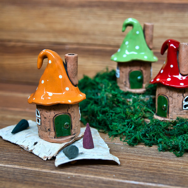 Drei skurrile Räucherhaus Keramik Zipfelmütze von Original Crottendorfer, jede mit einem hohen orangefarbenen, grünen oder roten Dach und aus Keramik gefertigt, werden auf Holz zwischen grünem Moos und kleinen kegelförmigen Akzenten ausgestellt.