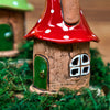 Das Räucherhaus Keramik Zipfelmütze von Original Crottendorfer ist ein liebevoll gestaltetes Deko-Haus mit rot-weißem Pilzdach, grüner Tür und weißem Fenster, das auf grünem Moos platziert ist.