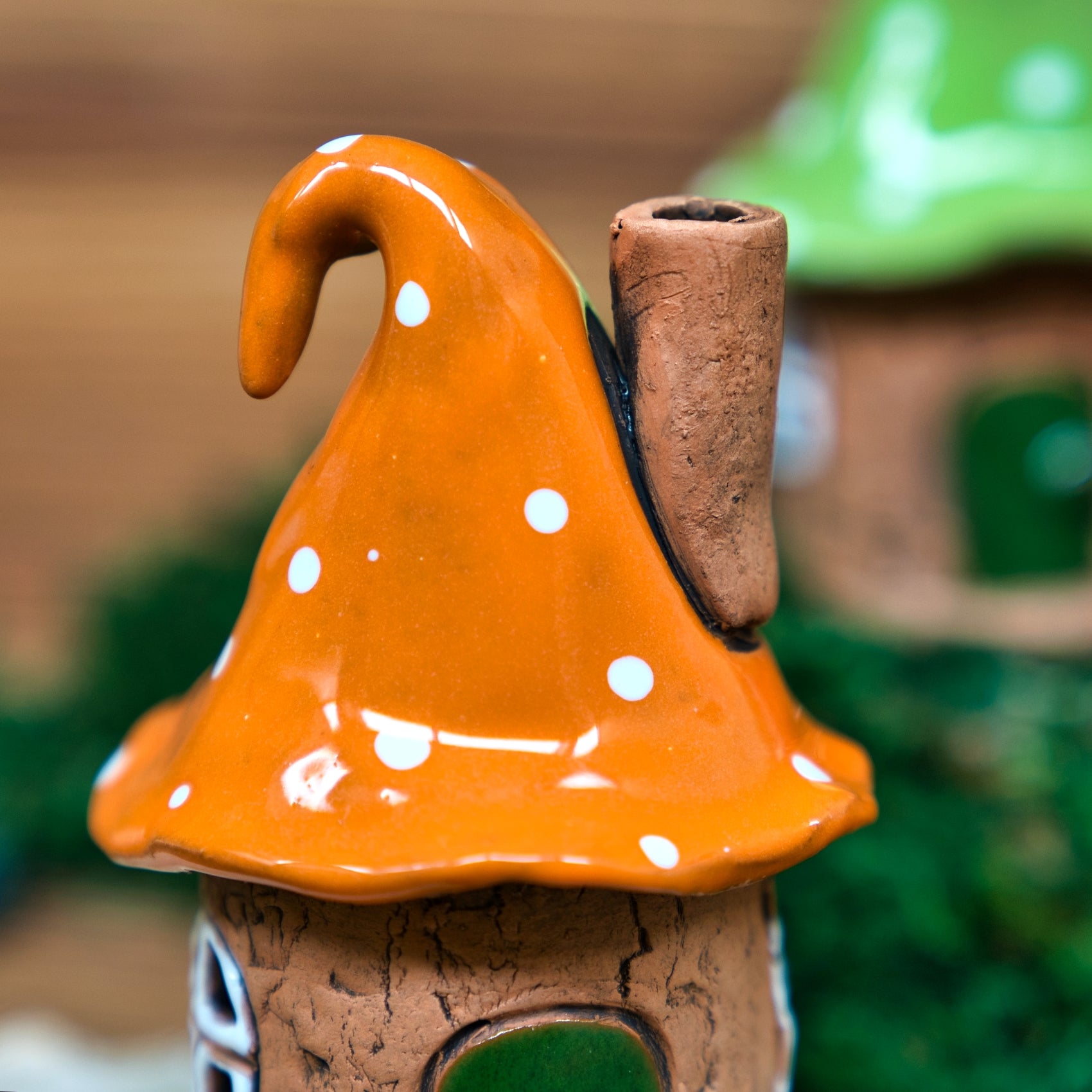 Eine Nahaufnahme des Original Crottendorfer Räucherhauses Keramik Zipfelmütze mit orangefarbenem, gepunktetem Pilzkopfdach und einem Schornstein. Im unscharfen Hintergrund befindet sich ein weiteres charmantes Haus mit einem grünen Dach.
