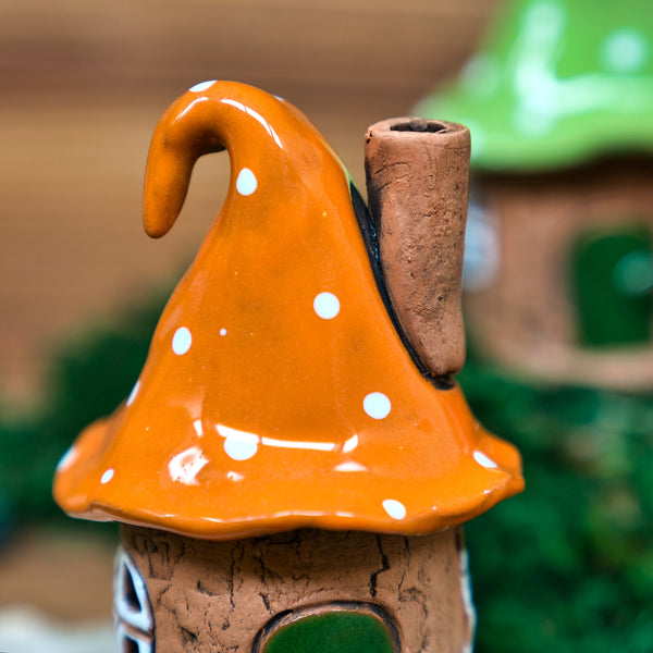Eine Nahaufnahme des Original Crottendorfer Räucherhauses Keramik Zipfelmütze mit orangefarbenem, gepunktetem Pilzkopfdach und einem Schornstein. Im unscharfen Hintergrund befindet sich ein weiteres charmantes Haus mit einem grünen Dach.