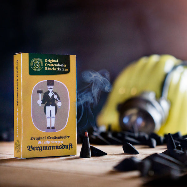 Neben einer brennenden Räucherkerze strömt duftender Weihrauch aus einer Schachtel „Original Crottendorfer Räucherkerzen Nostalgie Edition Bergmannsduft“ in die Luft. Weitere Räucherkerzen sind überall verstreut, während im Hintergrund sanft ein gelber Bergmannshelm verschwimmt.