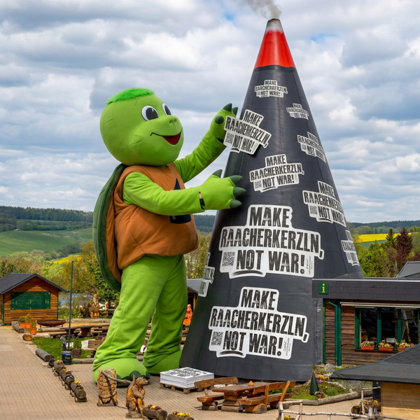 Ein riesiges Schildkrötenmaskottchen steht neben einem großen Kegel mit dem Aufkleber "Mach Raacherkerzln, nicht Krieg!" von Original Crottendorfer und feiert erzgebirgische Tradition inmitten von grünen Hügeln und Holzhäusern im Freien.
