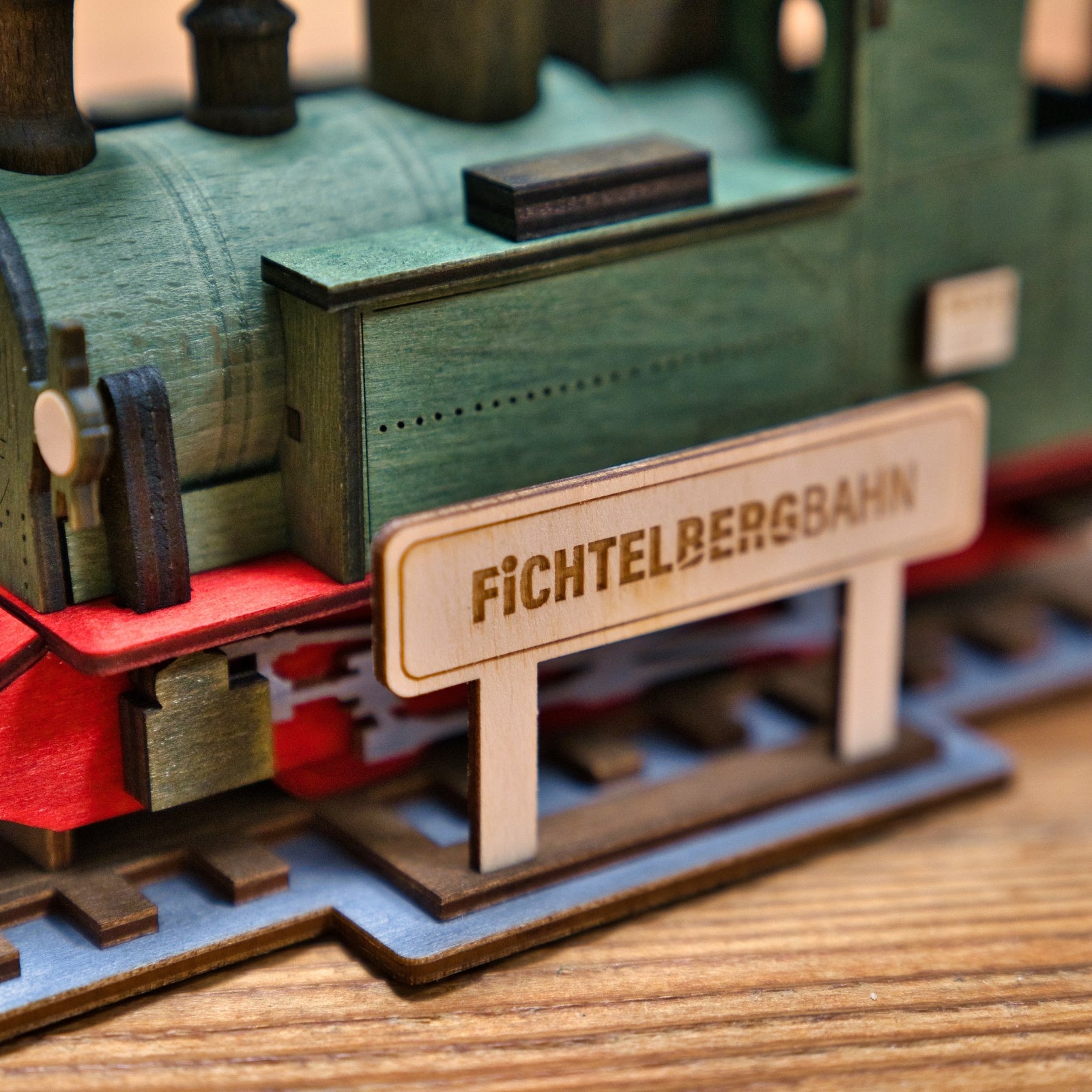 Eine Nahaufnahme der Lenk & Sohn Räucherlok Fichtelbergbahn grün zeigt ihr grün-rotes Design mit einem kleinen Holzschild. Dieses detaillierte Holzmodell fängt den Charme einer Dampflok-Abenteuer-Szene ein.