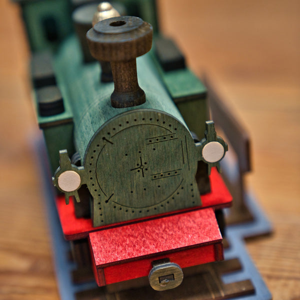 Nahaufnahme der Räucherlok Fichtelbergbahn grün von Lenk & Sohn, einer grünen Spielzeugeisenbahnlok mit roten Akzenten auf grauem Gleis. Dieses Holzmodell ist ideal für Liebhaber. Die Vorderseite ist mit detaillierten runden und rechteckigen Motiven versehen.