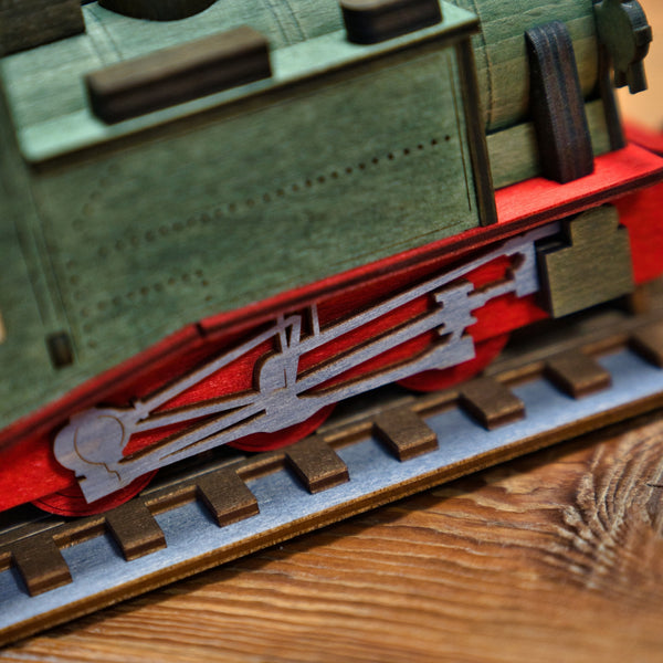 Nahaufnahme der Räucherlok Fichtelbergbahn grün von Lenk & Sohn mit detaillierten grünen und roten Teilen, Antriebsstangen und Rädern auf der Schiene, die auf einer Holzoberfläche ihren einzigartigen Charme entfalten.