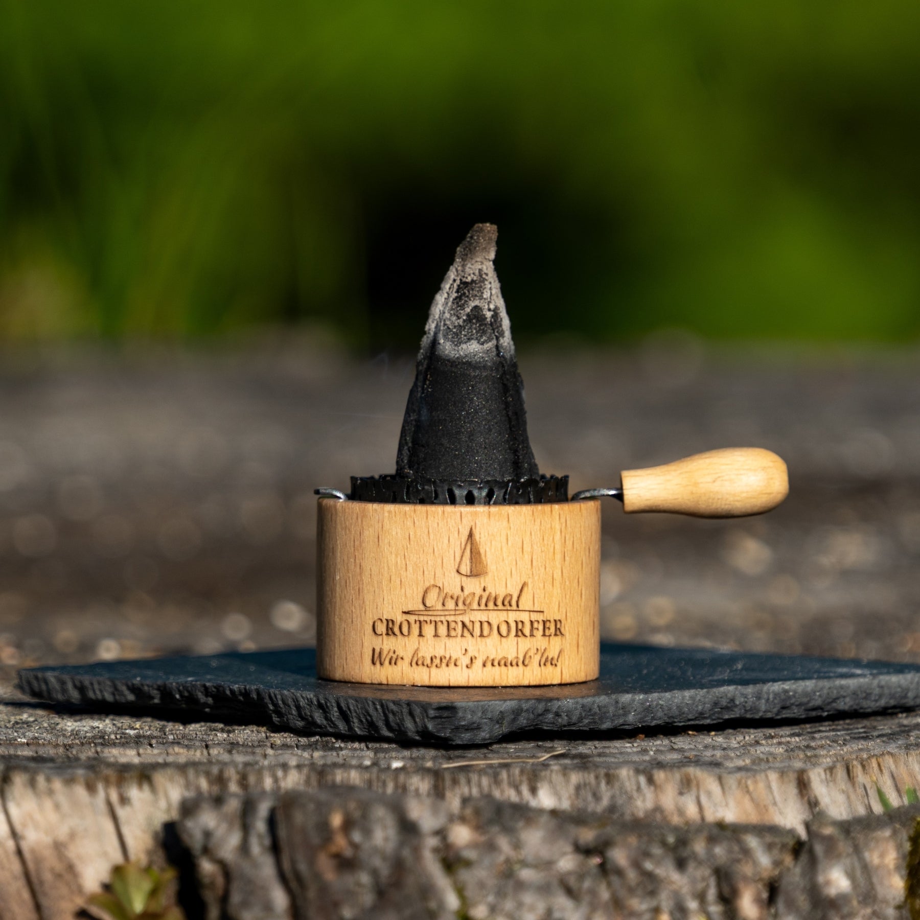 Das Räuchertöpfchen von Original Crottendorfer Räucherkerzen, ein Holzwerkzeug mit deutscher Gravur, hält einen großen schwarzen Kegel auf einer runden Schieferplatte im Freien, die auf Holz mit einem verschwommenen grünen Hintergrund steht.