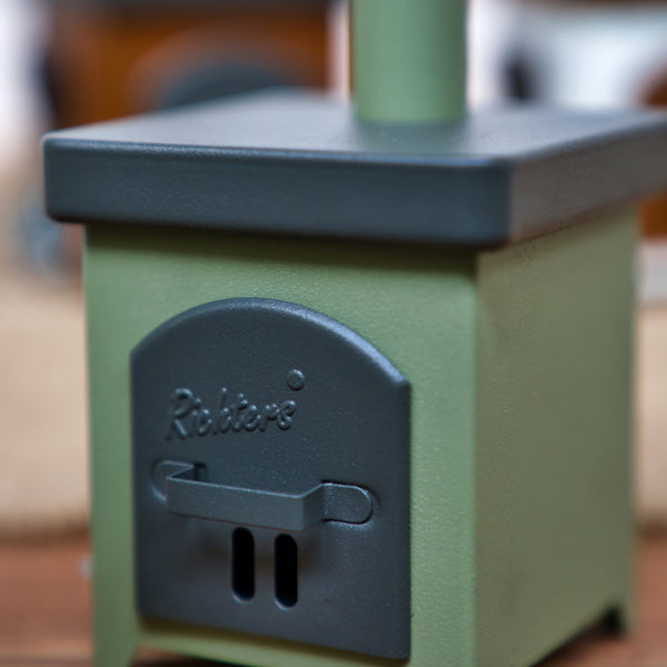 Eine Nahaufnahme des Räucherofens Hüttenofen, eines kleinen grünen Metallofens von Hartmut Richter mit schwarzer Tür und geprägtem Brandzeichen, der charmant auf einer Holzfläche im Innenbereich steht.