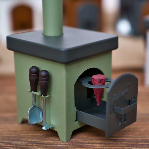 Der Räucherofen Hüttenofen von Hartmut Richter ist ein kleiner grüner Räucherofen, der einem Holzofen nachempfunden ist, mit Miniaturutensilien an der Seite und einem roten Kegel in der offenen Tür, der auf einer Holzfläche steht.