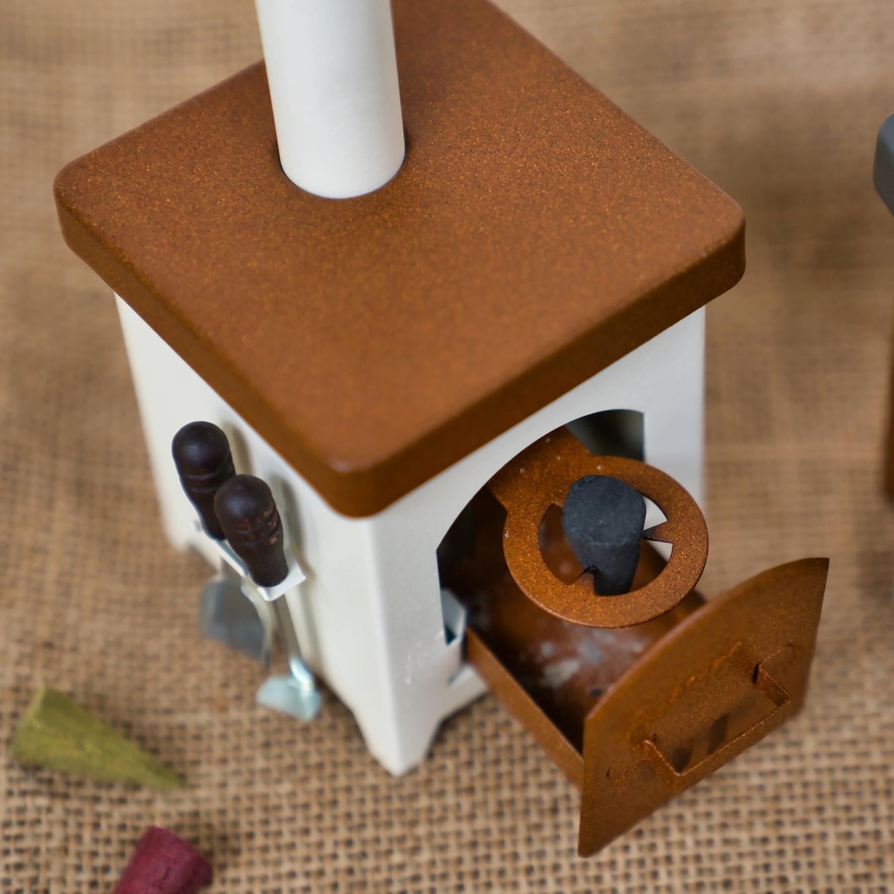 Ein kleiner Hartmut Richter Räucherofen Hüttenofen in Weiß und Rost, der eine Pfeife, eine offene Tür, kleine Utensilien und einen Aschekasten zeigt, liegt auf Sackleinen. In der Nähe sind bunte Kreidestücke verstreut.