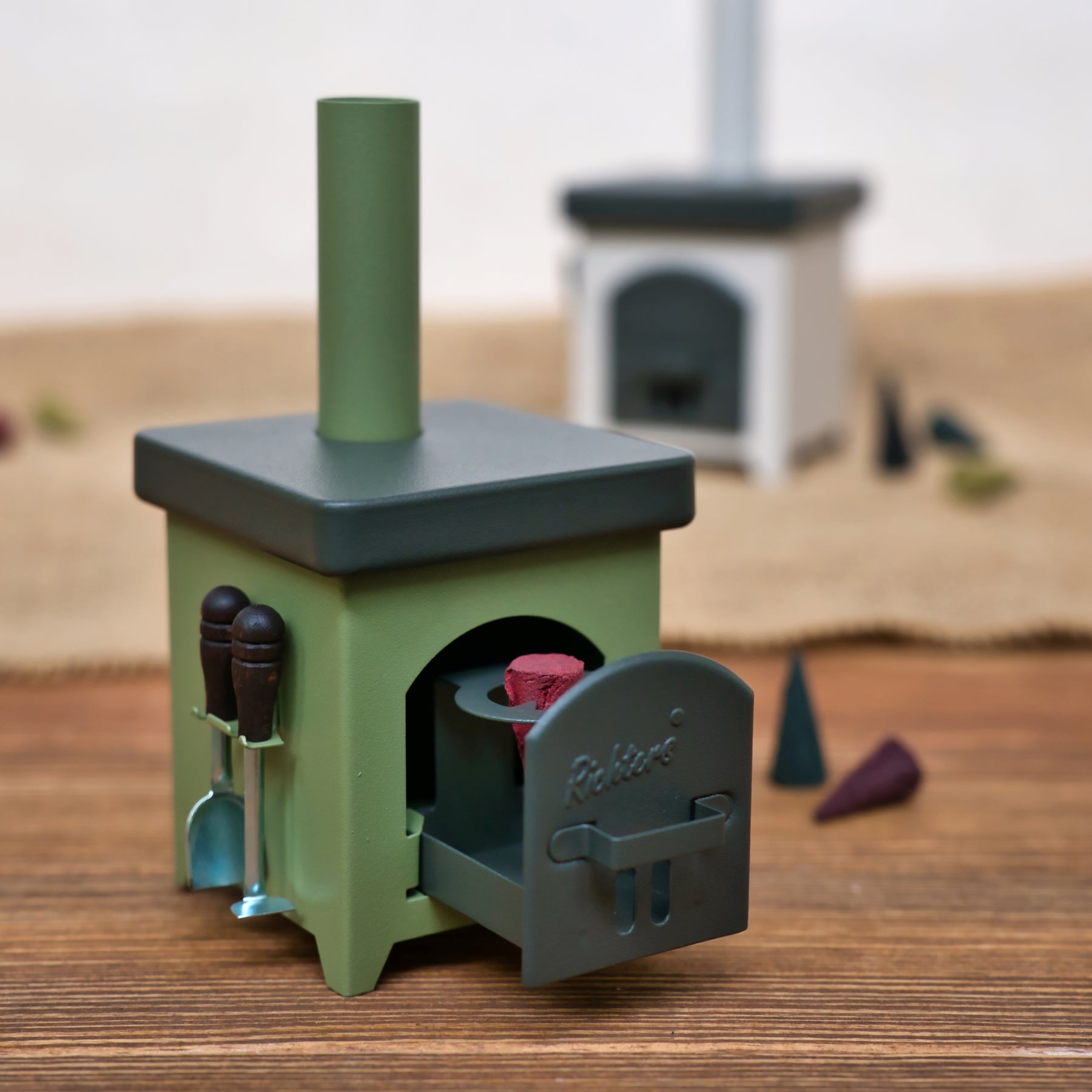 Ein kleiner, grüner Hartmut Richter Räucherofen Hüttenofen mit offener Tür und winzigen Werkzeugen an der Seite steht auf einer Holzfläche. Ein weiterer Miniatur-Hüttenofen ist im Hintergrund verschwommen zu erkennen.