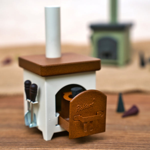 Der Hartmut Richter Räucherofen Hüttenofen, ein kleiner Spielzeug-Holzofen mit braunem Oberteil und weißem Korpus, ist mit einer offenen Tür und kleinen Utensilien an der Seite versehen. Ein weiterer unscharfer Ofen steht auf einer Holzfläche im Hintergrund.