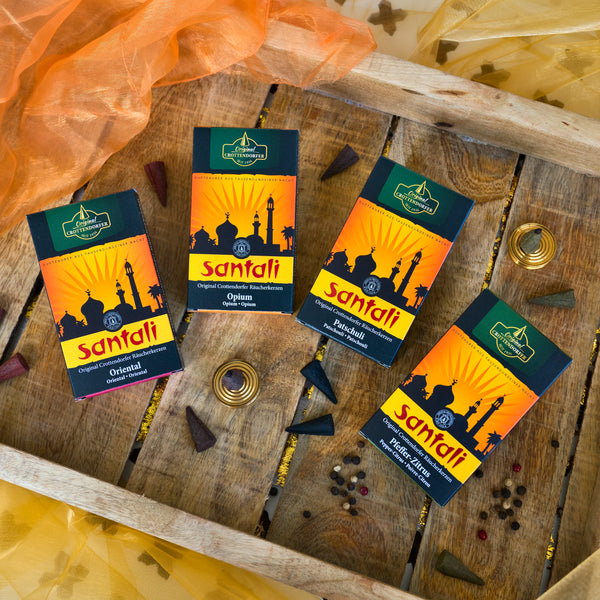 Schachteln mit Original Crottendorfer Räucherkerzen Patschuli werden auf einem Holztablett mit Räucherkegeln, Gewürzen, Siam Benzoe Harz und orange-gelbem Stoff für eine aphrodisierende Wirkung präsentiert.