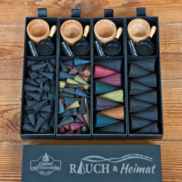 Vier „RAUCH & Heimat“-Sets von Original Crottendorfer Räucherkerzen, die jeweils kleine Holzschälchen, schwarze Metallschaufeln sowie bunte Räucherkegel und -stäbchen enthalten, sind ordentlich auf einer Holzunterlage angeordnet. Auf dem Deckel der Schachtel prangt der Schriftzug „Rauch & Heimat“, was diese Geschenksets zum perfekten Geschenk für Liebhaber hochwertiger Räucheraccessoires macht.