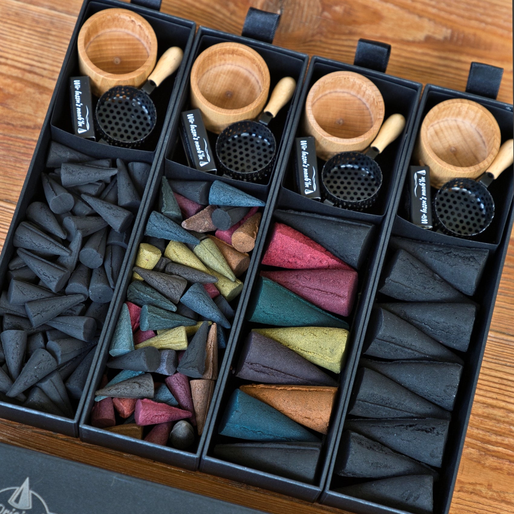 Eine stilvolle Schubbox „RAUCH & Heimat“ von Original Crottendorfer Räucherkerzen mit vier Fächern, gefüllt mit Räucherkegeln und -stäbchen in verschiedenen Formen und Farben, ergänzt durch Räucherhalter aus Holz und schwarze Netzunterlagen – alles wunderschön auf einem Holztisch präsentiert. Das perfekte Geschenk für alle, die edle Räucheraccessoires schätzen.