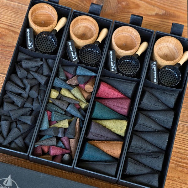 Eine stilvolle Schubbox „RAUCH & Heimat“ von Original Crottendorfer Räucherkerzen mit vier Fächern, gefüllt mit Räucherkegeln und -stäbchen in verschiedenen Formen und Farben, ergänzt durch Räucherhalter aus Holz und schwarze Netzunterlagen – alles wunderschön auf einem Holztisch präsentiert. Das perfekte Geschenk für alle, die edle Räucheraccessoires schätzen.