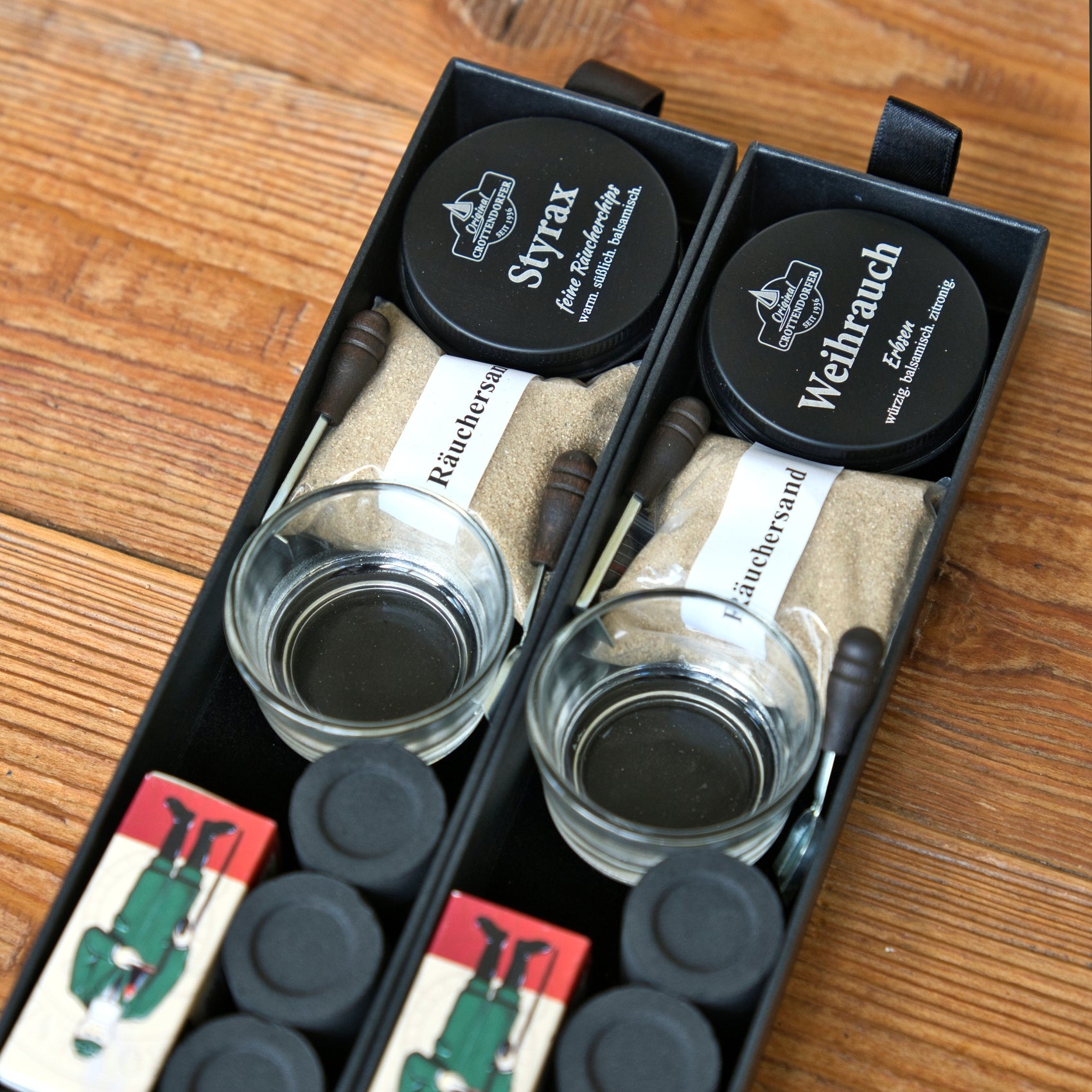 Auf einer Holzunterlage sind zwei „Schubbox RAUCH & Seele“-Sets von Original Crottendorfer Räucherkerzen ausgestellt, die jeweils ein Glas, Sand, zwei Behälter mit schwarzen, auf Deutsch beschrifteten Deckeln, Holzstäbchen und eine Streichholzschachtel mit der Abbildung eines Soldaten enthalten – alles, was man für die grundlegenden Räucherutensilien benötigt.