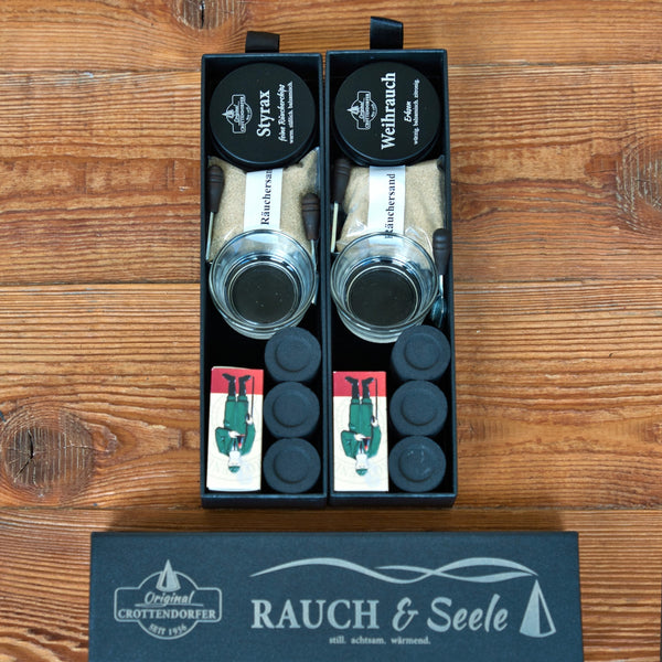 Zwei schwarze „Schubbox RAUCH & Seele“-Geschenkboxen von Original Crottendorfer Räucherkerzen enthalten Gläser, Räucherkegel, Streichholzschachteln und Glasbehälter. Die Schachteln sind mit „Styx“ und „Weihrauch“ beschriftet, während auf dem geschlossenen Deckel unten der Name „RAUCH & Seele“ zu sehen ist. Alle Räucherutensilien sind ordentlich auf einer Holzunterlage angeordnet.