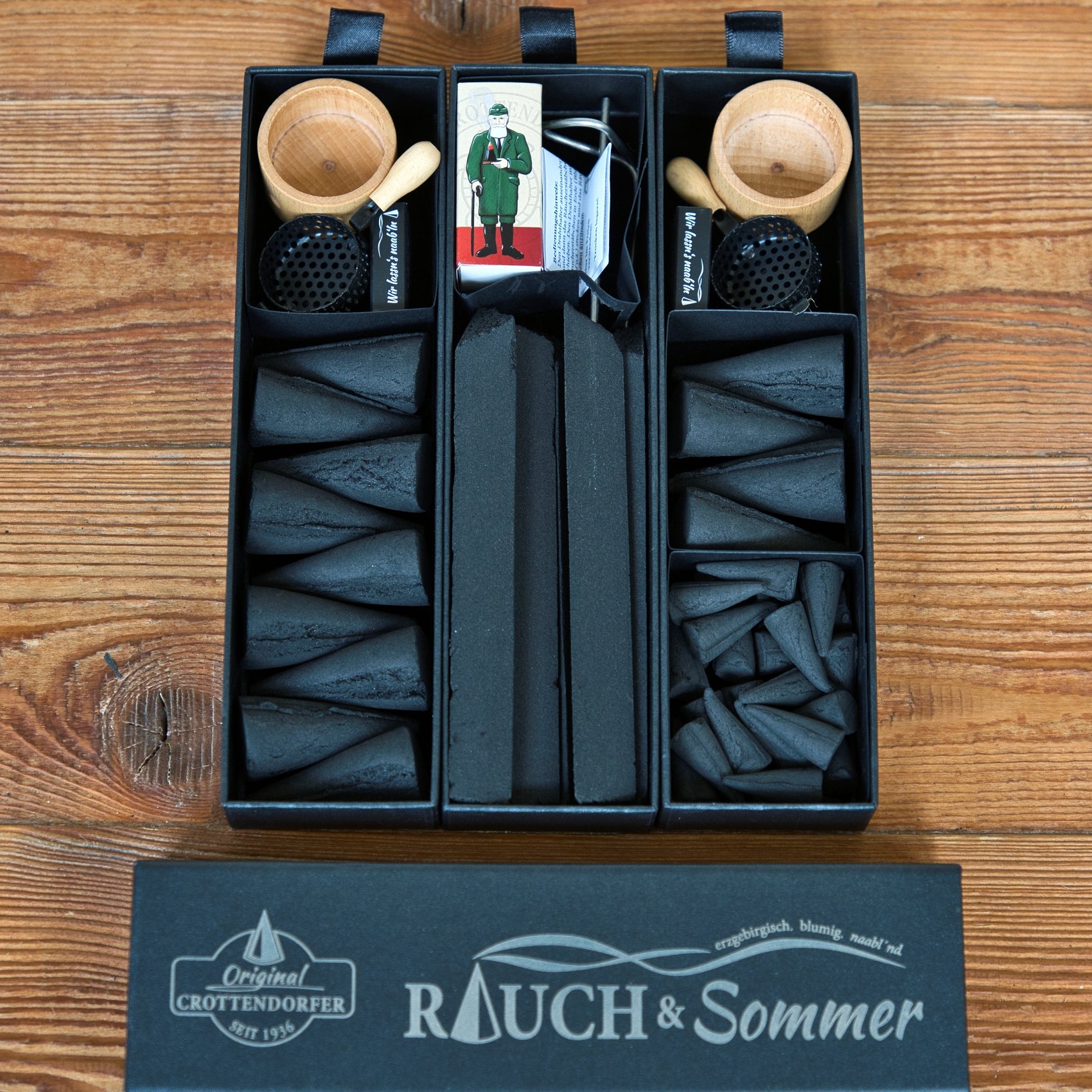 Die Schubbox „RAUCH & Sommer“ von Original Crottendorfer Räucherkerzen enthält drei Schalen, gefüllt mit Räucherkegeln, Räucherstäbchen, Räucherkerzen, kleinen Holzpfeifen, einer Streichholzschachtel und Metallgittern für einen wirksamen Mückenschutz. Das Set wird auf einer Holzunterlage präsentiert, auf deren Unterseite „RAUCH & Sommer“ steht.