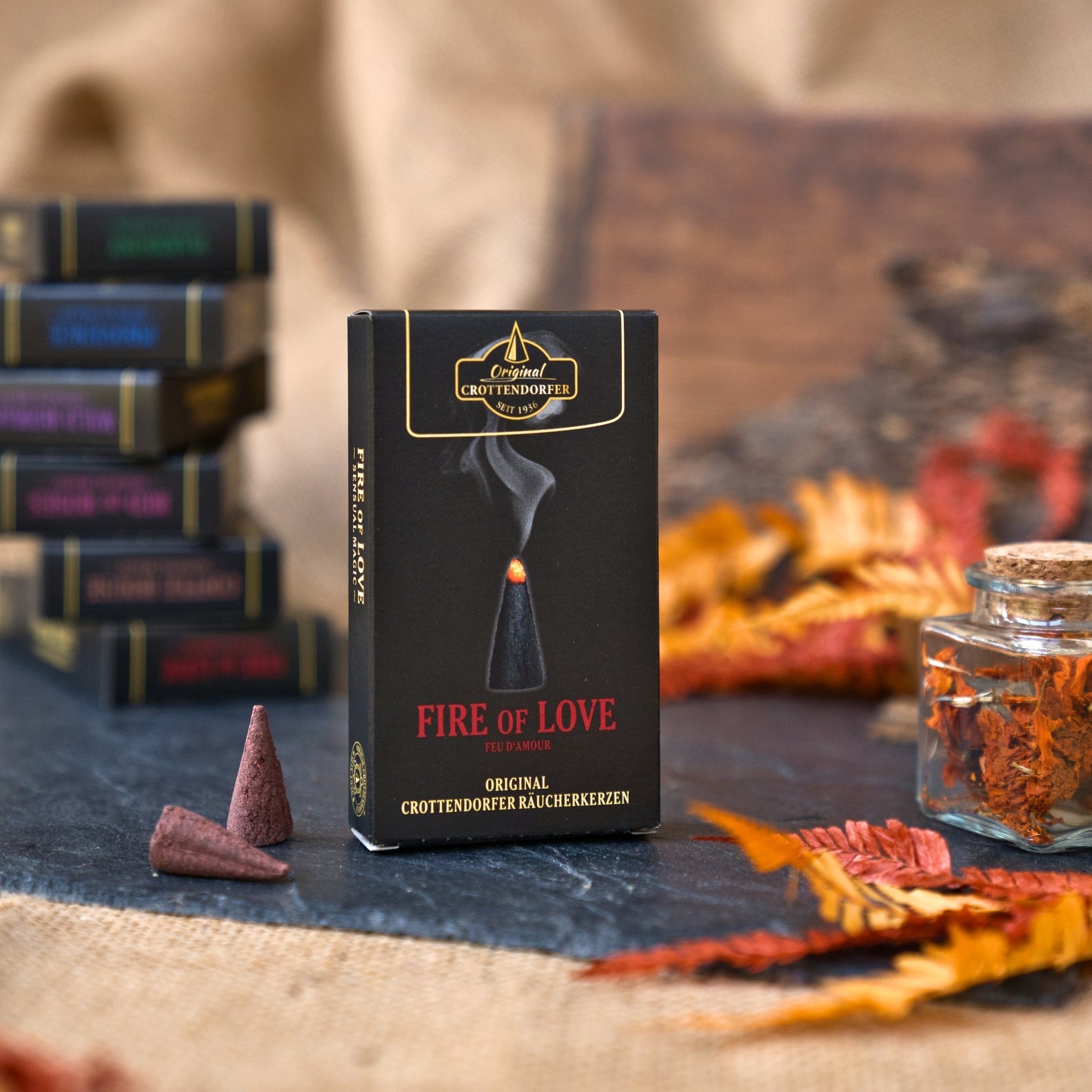 Eine Schachtel mit der Aufschrift „Original Crottendorfer Räucherkerzen Fire of Love“ steht aufrecht da, daneben liegt eine Räucherkegel. Der warme Duft von „süße Vanille“ liegt in der Luft und harmoniert mit dem Herbstlaub, einem mit getrockneten Kräutern gefüllten Glasgefäß und den verschwommenen Räucherkisten, die im Hintergrund verblassen.