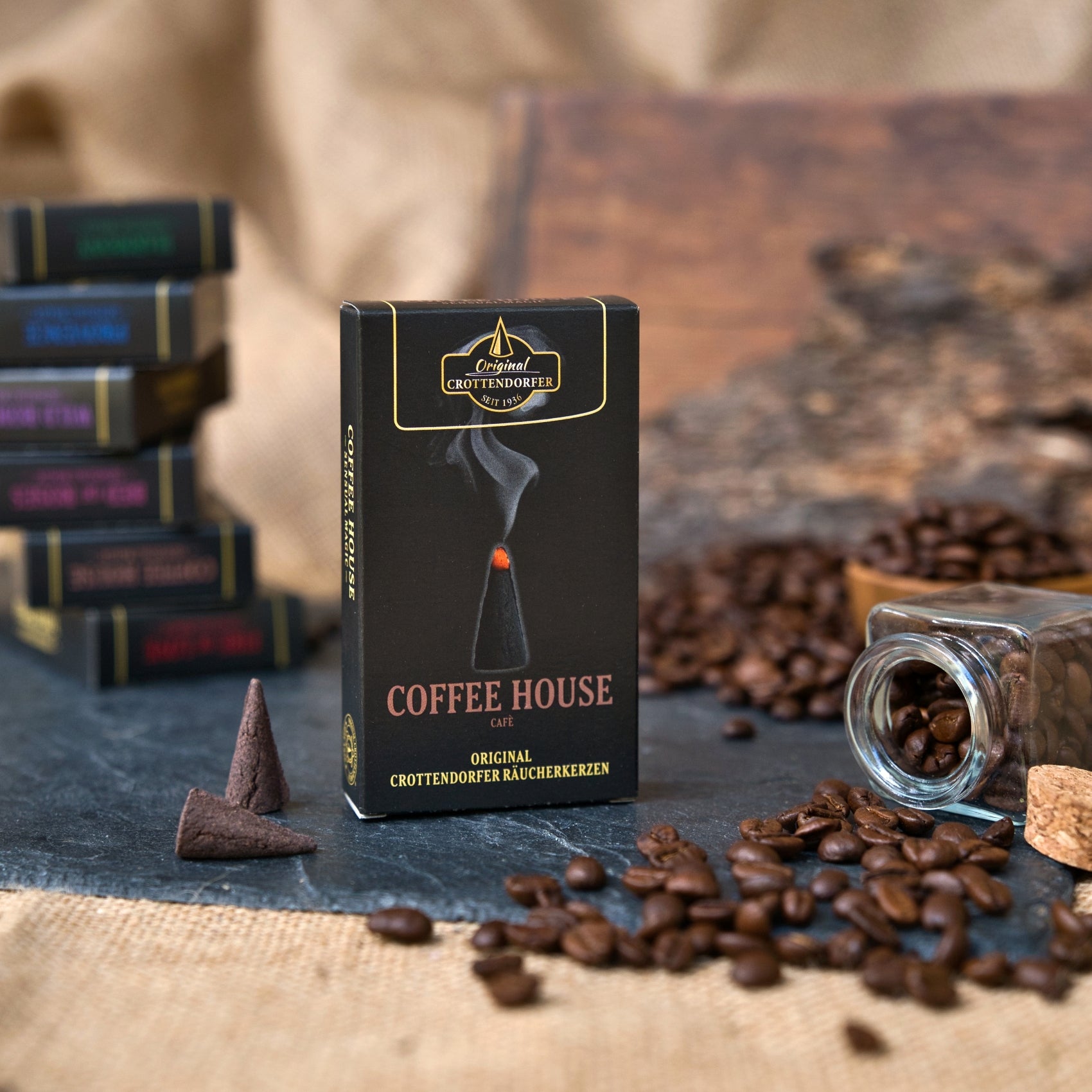 Eine Schachtel mit Original Crottendorfer Räucherkerzen „Coffee House“ steht inmitten von verstreuten Kaffeebohnen, einem umgestürzten Glasgefäß, weiteren Räucherkerzen und verschwommenen Schachteln im Hintergrund auf einer rustikalen Unterlage und verströmt den einladenden Duft von „Kaffeehaus“.