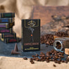 Eine Schachtel mit Original Crottendorfer Räucherkerzen „Coffee House“ steht inmitten von verstreuten Kaffeebohnen, einem umgestürzten Glasgefäß, weiteren Räucherkerzen und verschwommenen Schachteln im Hintergrund auf einer rustikalen Unterlage und verströmt den einladenden Duft von „Kaffeehaus“.