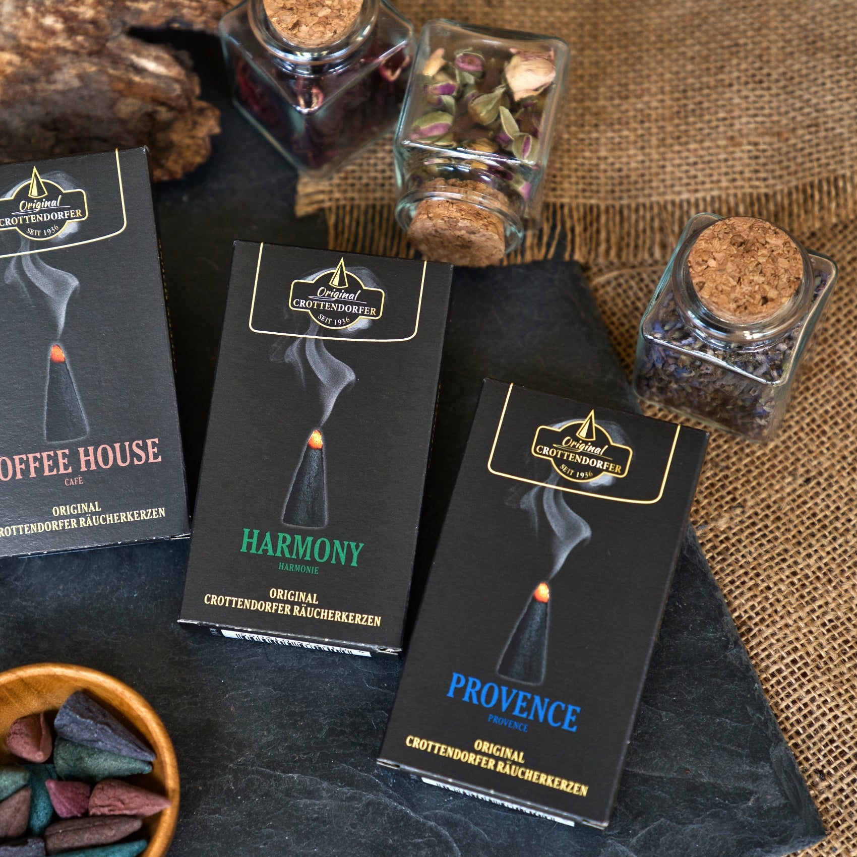 Drei Schachteln mit Original Crottendorfer Räucherkerzen – „Coffee House“, „Harmony“ und „Provence“ – sind auf einer Schieferplatte angeordnet, umgeben von kleinen Gläsern mit getrockneten Kräutern und Jute. Jede Sorte erfüllt die Luft mit ihrem unverwechselbaren herbstlichen Duft und zaubert mit jedem holzig-rauchigen Aroma eine gemütliche Herbststimmung herbei.