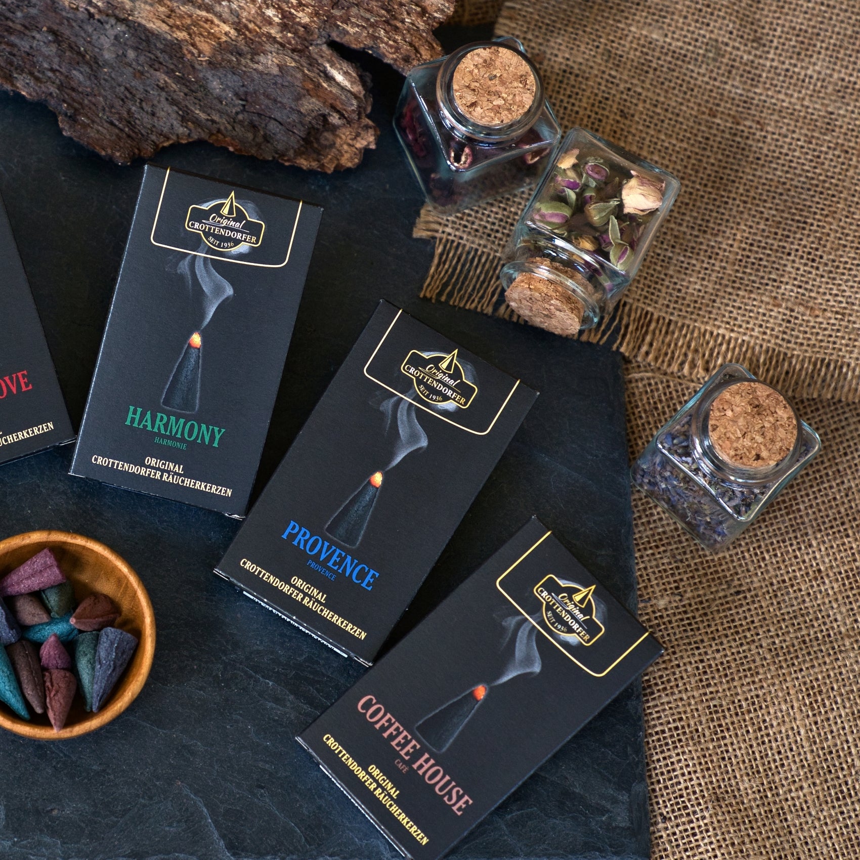 Drei schwarze Räucherkerzenschachteln von Original Crottendorfer Räucherkerzen – mit den Sorten „Harmony“, „Provence“ und „Coffee House“ – sind auf einer dunklen Unterlage neben Gläsern mit getrockneten Blumen angeordnet. Braunes Sackleinen und ein Stück Holz bilden den Hintergrund, während farbige Räucherkegel einen romantischen Duft verströmen, der an frische Gartennelken und französischen Honig erinnert – das charakteristische Aroma der Räucherkerzen „Provence“.