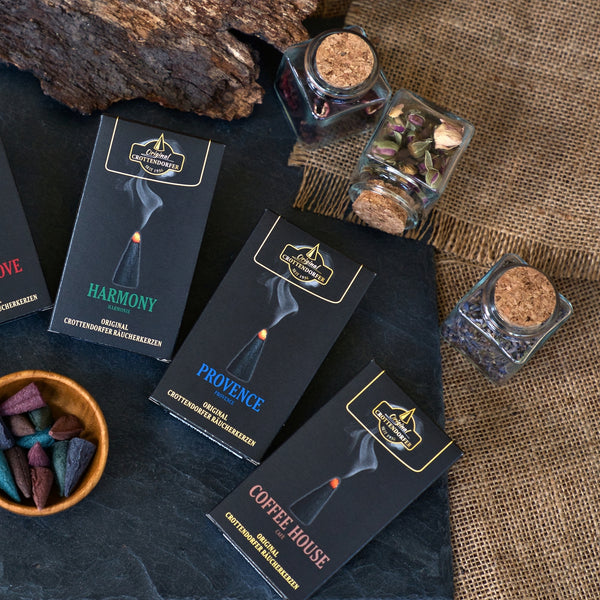 Drei schwarze Räucherkerzenschachteln von Original Crottendorfer Räucherkerzen – mit den Sorten „Harmony“, „Provence“ und „Coffee House“ – sind auf einer dunklen Unterlage neben Gläsern mit getrockneten Blumen angeordnet. Braunes Sackleinen und ein Stück Holz bilden den Hintergrund, während farbige Räucherkegel einen romantischen Duft verströmen, der an frische Gartennelken und französischen Honig erinnert – das charakteristische Aroma der Räucherkerzen „Provence“.