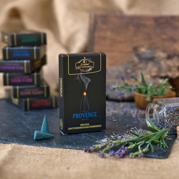 Eine Schachtel mit der Aufschrift „Räucherkerzen Provence“ von Original Crottendorfer Räucherkerzen steht aufrecht, davor liegt eine einzelne brennende Räucherkegel. Im Hintergrund liegen getrockneter Lavendel, ein kleines Glas und gestapelte Räucherkisten auf einer strukturierten Oberfläche und schaffen eine Atmosphäre voller romantischer Düfte und feiner Noten von französischem Honig.