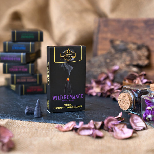 Auf einer dunklen Fläche steht eine schwarze Schachtel mit der Aufschrift „Räucherkerzen Wild Romance“ von Original Crottendorfer Räucherkerzen, umgeben von Räucherkegeln, Gläsern, getrockneten Blütenblättern und gestapelten Räucherkisten vor einem Hintergrund aus Sackleinen. Die Szene weckt den Duft des Mittelmeerraums, wobei sich Noten von frischen Feigen und zarte Akzente von Weihrauch in der Luft vermischen.