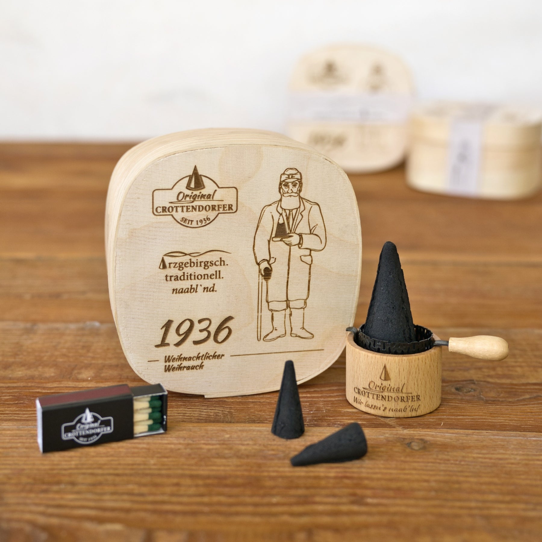 Ein reizvolles Geschenkset von Original Crottendorfer Räucherkerzen wird auf einer Holzunterlage präsentiert. Es umfasst die „Spanschachtel 1936“ – eine mit der Abbildung eines Mannes verzierte Holzschachtel – sowie einen kleinen Holzhalter mit schwarzen Räucherkegeln als ideales Räucherzubehör und eine dazugehörige Streichholzschachtel.
