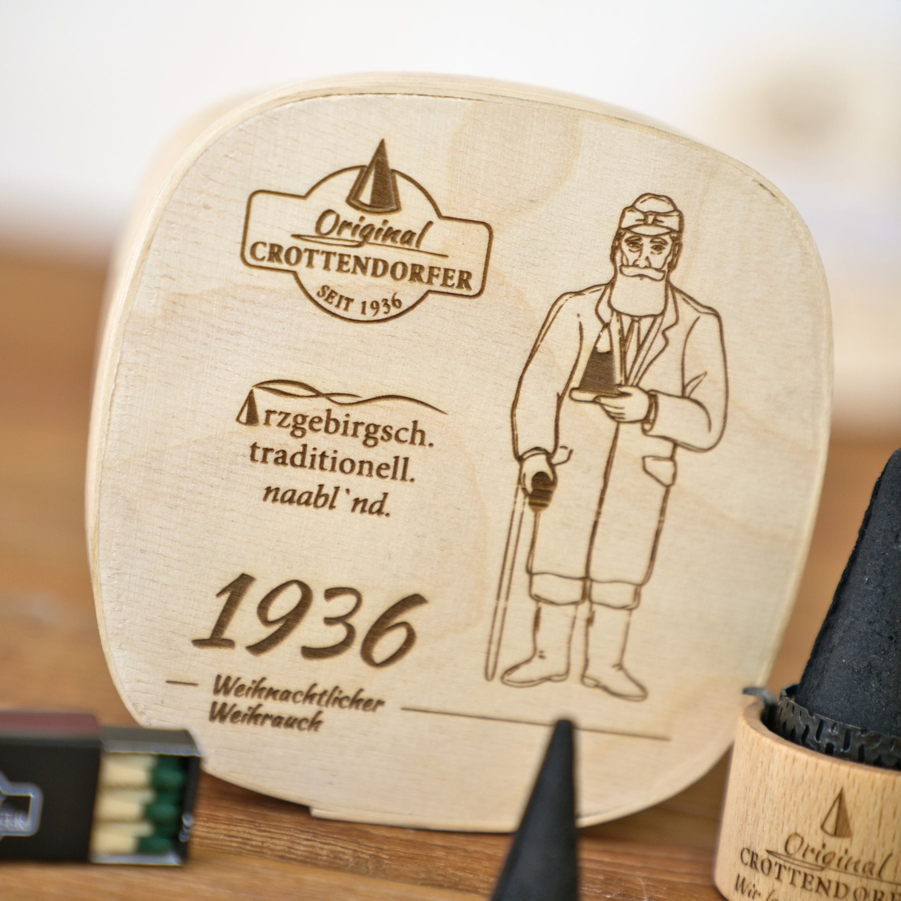Die „Spanschachtel 1936“ von Original Crottendorfer Räucherkerzen verfügt über eine Holzplakette, auf der ein bärtiger Mann in einem Mantel mit einem Spazierstock eingraviert ist, begleitet von einem deutschen Text und der Jahreszahl 1936. Zusammen mit den kegelförmigen schwarzen Objekten ist dieses Set ein ideales Geschenkset für Sammler traditioneller Spanschachteln oder Liebhaber einzigartiger Räucherzubehörteile.
