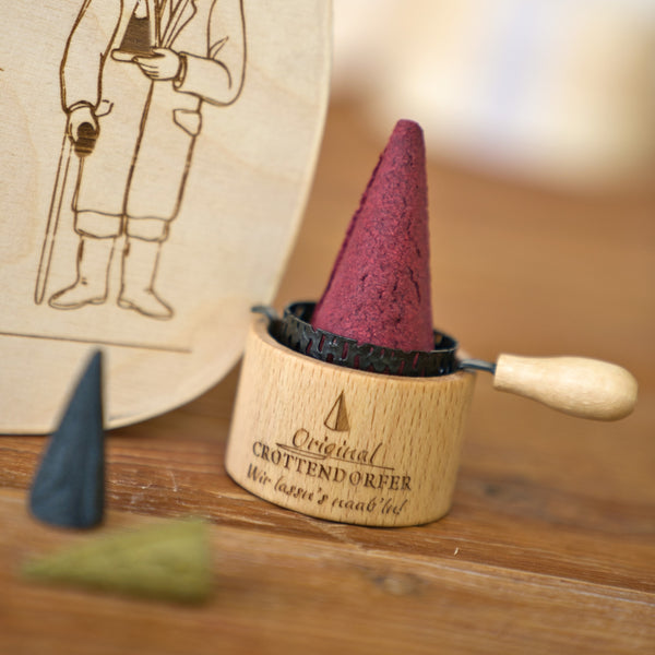 Ein roter Räucherkegel von Original Crottendorfer Räucherkerzen steht in einem Holzhalter mit Griff; auf dem Tisch daneben liegen zwei weitere Kegel, einer in Schwarz und einer in Grün. Im Hintergrund rundet ein Teil der „Spanschachtel 1936“ – eine wunderschön illustrierte Holzschachtel – dieses bezaubernde Räucherzubehör-Arrangement ab, das sich ideal als Teil eines liebevoll zusammengestellten Geschenksets eignet oder zusammen mit anderen traditionellen Stücken präsentiert werden kann.