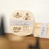 Eine quadratische Spanschachtel aus Holz von „Original Crottendorfer Räucherkerzen“ aus dem Jahr 1936 mit der Abbildung eines bärtigen Mannes und dem Schriftzug „Original Crottendorfer 1936“, steht auf einer weißen Unterlage, die mit einem Zierband umwickelt ist – ein ideales Geschenkset für Liebhaber traditionellen Räucherzubehörs.