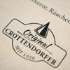 Nahaufnahme eines beigen Stoffetiketts auf dem Stoffbeutel „Raacherkerzl Definition“ oder „Bitte nicht schubsen ...“ von Original Crottendorfer, das das schwarze Logo „Original Crottendorfer Seit 1936“ mit Kegelmotiv und Verweisen auf die erzgebirgische Tradition zeigt.