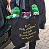 Eine Person hält einen Original Crottendorfer Stoffbeutel („Raacherkerzl Definition“ oder „Bitte nicht schubsen ...“) in der Hand, der zwei grüne Plüschfrosche und Crottendorfer Räucherkerzen enthält; Im Hintergrund sind Kopfsteinpflaster und ein Auto zu sehen.