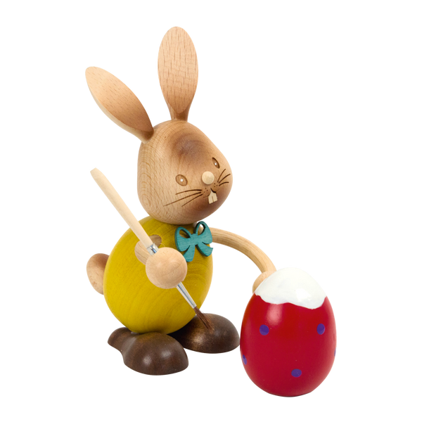 Der Stupsi Hase Eiermaler von der Drechslerei Kuhnert ist eine hölzerne Hasenfigur mit gelbem Körper, grüner Fliege und braunen Füßen, die einen Pinsel hält, während sie ein rotes Ei mit blauen Punkten und weißem Deckel verziert.