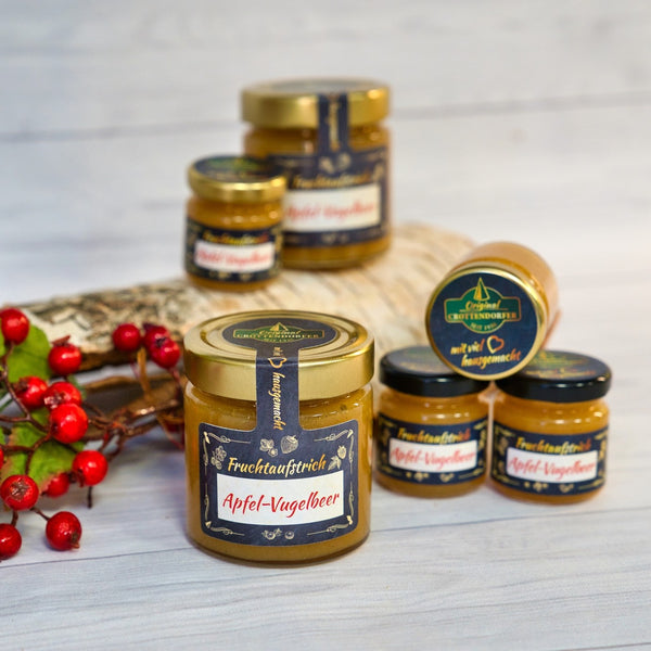 Auf einer Holzunterlage sind Gläser mit „Original Crottendorfer Hausgemachter Fruchtaufstrich“ (50 g) angeordnet, umrahmt von roten Beeren und Blättern als Dekoration. Diese vegane Köstlichkeit wird vor einem sanften, unscharfen Hintergrund präsentiert.