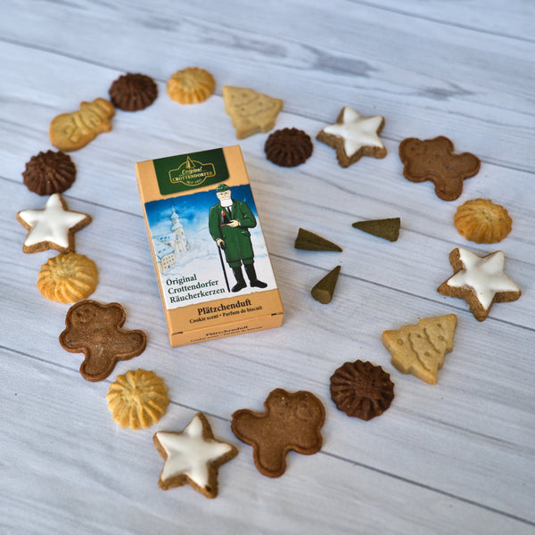 Eine Schachtel „Räucherkerzen Plätzchenduft“ von Original Crottendorfer Räucherkerzen ist umgeben von Plätzchen, darunter sternförmige Lebkuchenmänner und runde Butterkekse, die herzförmig auf einer hellen Holzoberfläche angeordnet sind.
