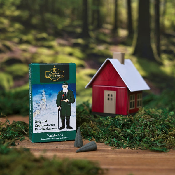 Eine Schachtel Original Crottendorfer Räucherkerzen Räucherkerzen Waldmoos steht auf Moos neben zwei Zapfen und einem kleinen roten Modellhaus. Der Hintergrund ist waldig und erinnert an den reichen Duft von Waldmoos.