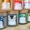 Eine Nahaufnahme von Candle Factory Aromabadekissen "Männerbad" Duftkerzen in Milchglasgefäßen mit bunten Motiven und Holzdeckeln, angeordnet auf einer hellen Fläche - perfekt für ein entspannendes Bad.