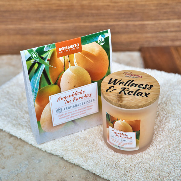 Eine Kerze im Glas und die dazu passende Dose mit Aprikosenmotiven und dem Branding "Augenblicke im Paradies" von Candle Factory ruhen auf einem weißen Handtuch - perfekt abgestimmt auf das Aromabadekissen "Augenblicke im Paradies" für echte Entspannung.