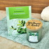 Ein grünes Päckchen mit der Aufschrift Aromabadekissen Erkältungszeit und eine Candle Factory Wellness-Duftkerze Erkältungszeit mit einem Holzdeckel mit der Aufschrift Wellness & Relax liegen auf einem hellblauen Handtuch, mit einem beigen Handtuch im Hintergrund.