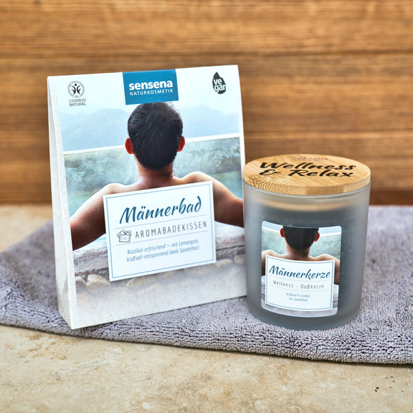Eine Dose Candle Factory Aromabadekissen "Männerbad" mit Sandelholz Duft und eine Wellness & Relax Männerkerze sitzen auf einem grauen Handtuch vor einem Holzhintergrund. Beide zeigen auf ihren Verpackungen einen Mann, der ein entspannendes Bad genießt.