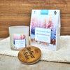 Die Candle Factory Wellnessduftkerze "Wintermärchen" aus Milchglas ruht auf einem weißen Handtuch neben einem Holzdeckel mit der Gravur Wellness & Relax und einem bunten Winterwald-Aromatherapie-Badesäckchen.