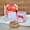 Das Aromabadekissen "Zauber der Rose" von Candle Factory mit einer rosafarbenen Kerze und einem runden Bambusdeckel mit der Gravur "Wellness & Relax" liegt auf einer strukturierten Oberfläche und erfüllt die Luft mit sanftem Rosenduft und ätherischen Ölen vor einem hölzernen Hintergrund.