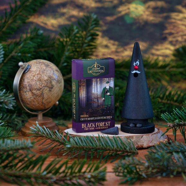 Ein kleiner Globus, eine Schachtel Original Crottendorfer Räucherkerzen Schwarzwald und ein schwarzer Kegelhalter mit Kulleraugen und roter Nase stehen auf einem Holz mit Tannenzweigen vor einer Waldkulisse.
