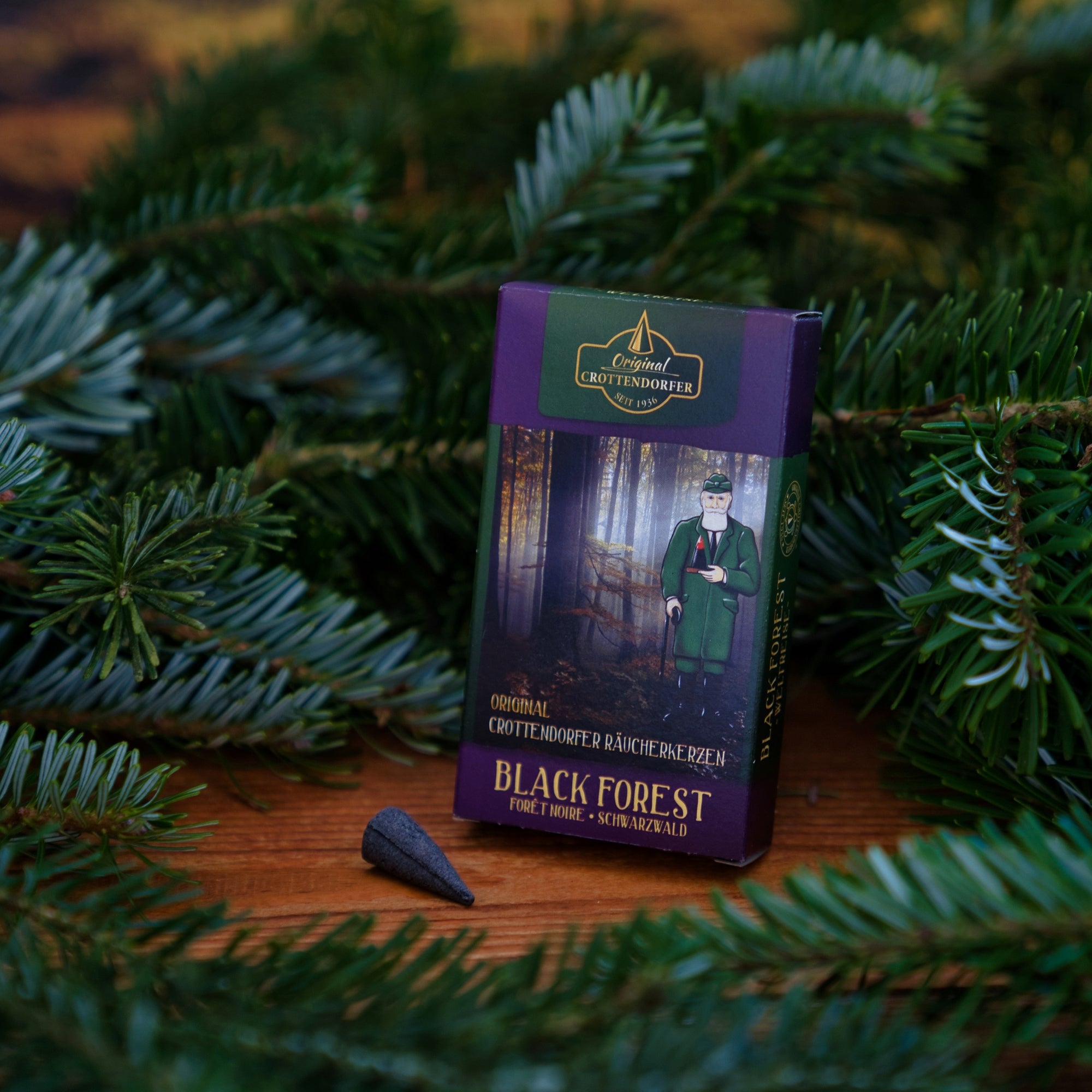 Eine Dose Original Crottendorfer Räucherkerzen Schwarzwald liegt auf einem Holz inmitten von Tannenzweigen. Auf der lila-grünen Schachtel ist ein Mann in Tracht abgebildet, der an den Schwarzwaldduft der Wildkräuter erinnert; davor steht ein Räucherkegel.