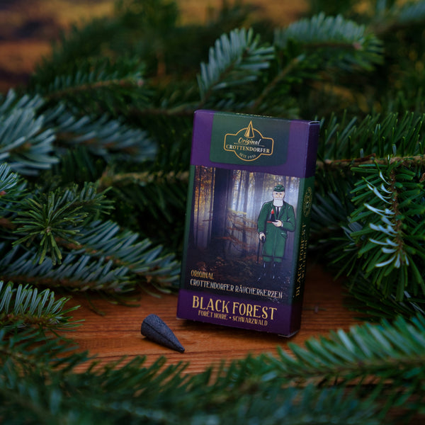 Eine Dose Original Crottendorfer Räucherkerzen Schwarzwald liegt auf einem Holz inmitten von Tannenzweigen. Auf der lila-grünen Schachtel ist ein Mann in Tracht abgebildet, der an den Schwarzwaldduft der Wildkräuter erinnert; davor steht ein Räucherkegel.